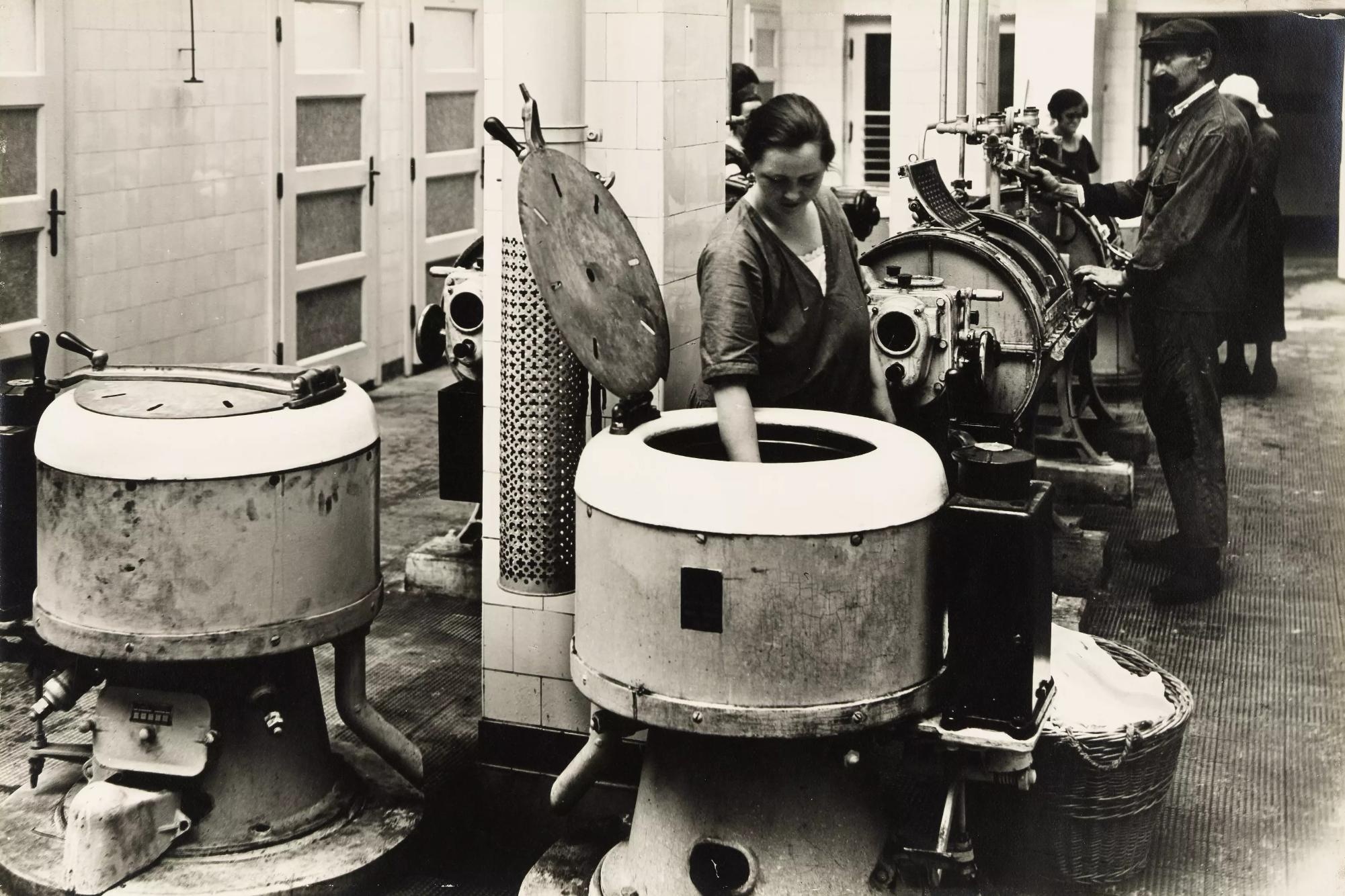 Eine Frau holt Wäsche aus dem Trog einer großen Waschtrommel, dahinter ein Mann und zwei Frauen an Waschgeräten. © © Wien Museum