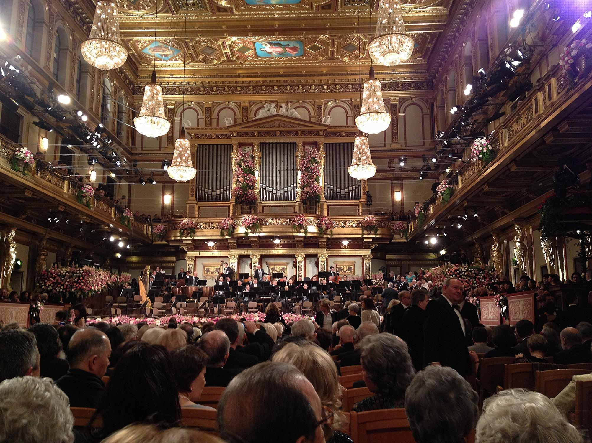 Das Bild zeigt Reihen von Zuschauer:innen beim Neujahrskonzert im großen Musikvereinssaal. Die Decke und die Logen sind mit goldenem Stuck und Malereien verziert. Im Hintergrund des Fotos befindet sich die Bühne mit den Musiker:innen. Von der Decke hängen sechs große Kronleuchter.  © Wikimedia, Michael Osmenda, Wiener Neujahrskonzert, 2013, CC-BY-2.0. 