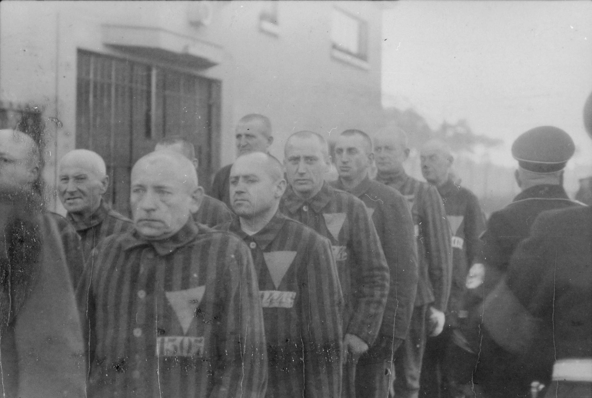 Häftlinge des KZ Sachsenhausen stehen vor einem Haus in der Reihe. Sie werden von Aufsehern bewacht. © National Archives, Heinrich Hoffmann, Häftlinge im Konzentrationslager Sachsenhausen, 1938.
