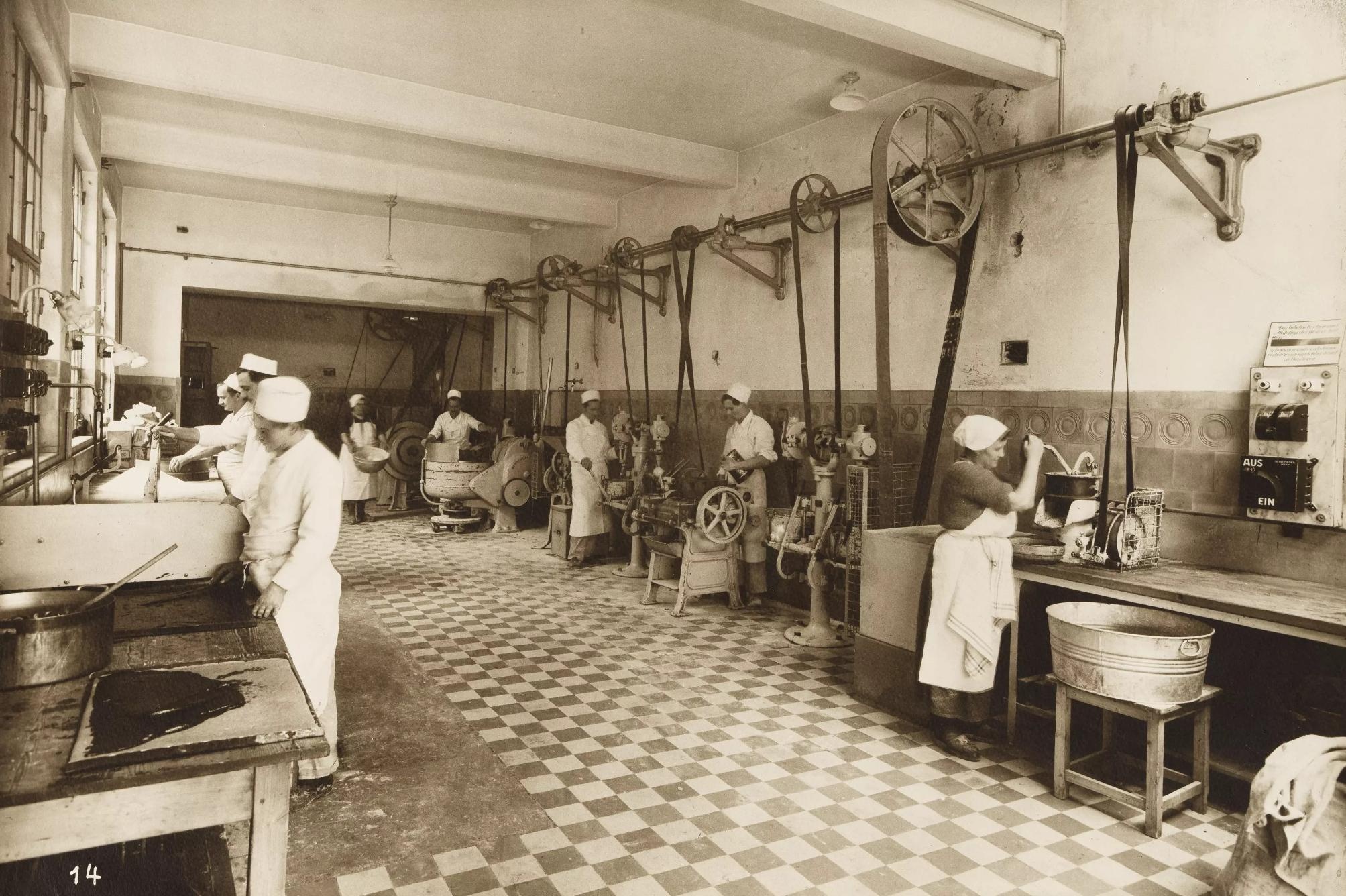 Frauen mit Schürze und Kopfbedeckung stehen an der linken und rechten Seite eines großen Raumes und kochen mit damals modernen Geräten.  © Wien Museum