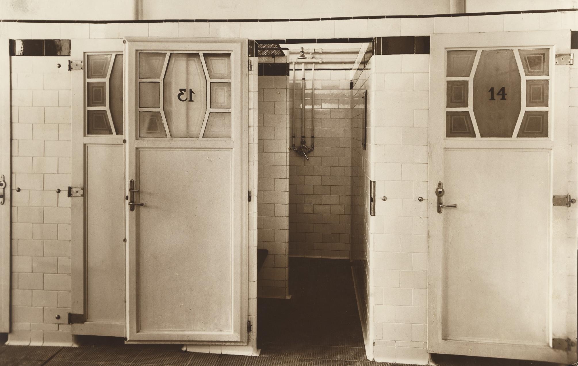 Das Foto zeigt weiß geflieste Wände und Gänge mit weißen Türen, die zu den Duschkabinen führen. In den Türen befindet sich oben ein Fenster mit einer Nummer, einmal „13“ und einmal „14“. Der Boden ist dunkel.  © Wien Museum, Städtisches Volksbad in der Ratschkygasse, 1926. 