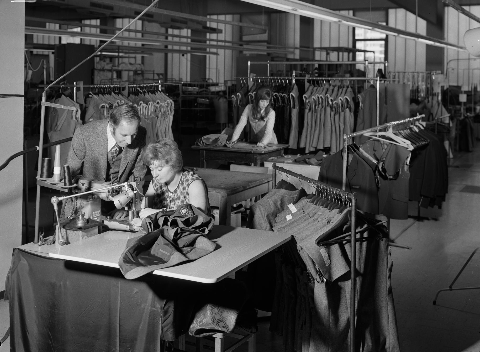 Eine Frau bedient eine Nähmaschine auf einem Schreibtisch in einer Näherei, ein Mann beugt sich über sie und sieht ihr bei der Arbeit zu. Rundherum Kleiderständer, im Hintergrund eine zweite Frau an einem Schreibtisch.  © ÖNB Bildarchiv