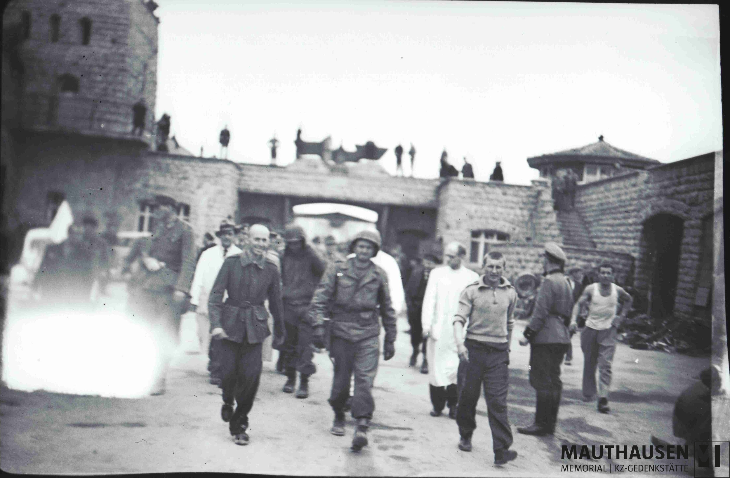 Das Schwarz-Wei&szlig;-Foto zeigt den Garagenhof des KZ Mauthausen am 5. Mai 1945. Im Hintergrund ist ein Tor zu sehen, &uuml;ber dem ein Reichsadler befestigt ist. In der Bildmitte befindet sich Albert Kosiek in Milit&auml;runiform und mit Helm, der zu Fu&szlig; den Hof durchquert. Au&szlig;erdem sind im Hof einige KZ-&Uuml;berlebende sowie Wachen der Wiener Feuerschutzpolizei in Uniform. Links im Bild befindet sich ein wei&szlig;er Fleck durch eine Besch&auml;digung der Kamera &copy; Mauthausen Memorial, Sammlung Antonio Garcia, S 4665, Fotograf: Francisco Boix, Albert Kosiek im Garagenhof, 5. Mai 1945, Signatur: 4.2.2.003.B3.