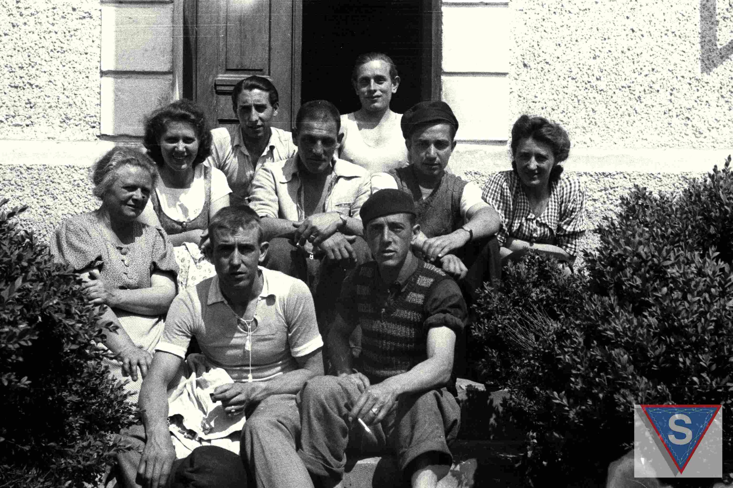 Das Schwarz-Wei&szlig;-Foto zeigt eine Gruppe von Menschen, die vor einem Haus auf der Treppe zur Eingangst&uuml;r sitzen. Ganz links sitzt Anna Pointner. Sie tr&auml;gt eine Bluse mit Kn&ouml;pfen und kurzen &Auml;rmeln. Ihre Haare sind hochgesteckt. In der Mitte der Gruppe sitzen sechs Spanier, die f&uuml;r die Kamera posieren. Einer davon l&auml;chelt, die anderen schauen ernst in die Kamera. Ein Mann ganz vorne h&auml;lt eine Zigarette in der Hand. Links und rechts der Spanier sitzt jeweils eine Tochter von Anna Pointner und l&auml;chelt in die Kamera. Im Vordergrund rahmen B&uuml;sche die Gruppe. &copy; Museu d'Hist&ograve;ria de Catalunya, Fons Amical de Mauthausen, Fotograf: Francisco Boix, Anna Pointner mit zwei T&ouml;chtern und sechs Spaniern vor ihrem Haus in Mauthausen, Mai 1945. 