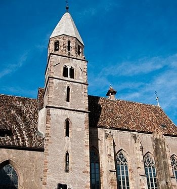 Die Kirche des Franziskanerklosters besteht aus graubraunen Steinmauern und einem Schindeldach mit verschiedenfarbigen Schindeln. Mittig im Foto befindet sich ein Kirchturm mit schmalen, spitz zulaufenden Fenstern. Die Spitze des Kirchturms besteht aus einem hellen Dach. Auch die Fenster des Kirchenschiffs laufen spitz zu und sind teilweise mit verschnörkelten Elementen verziert.  © Wikimedia, Bigmike.it, Franziskanerkloster in Bozen (CC-BY-SA-3.0). 