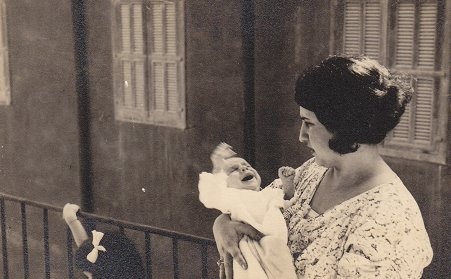Eine Mutter mit dunklen Haaren und angespanntem Gesichtsausdruck hält ein Baby im Arm. Das Baby schreit und strampelt. Links neben der Frau ist der Kopf eines Mädchens mit weißer Schleife im Haar zu sehen.  © Wikimedia, Mutter mit Baby, 1935. 