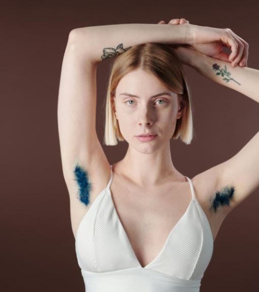 Eine Frau mit blondem Bob und Tattoos im weißen Top hebt die Arme nach oben, wodurch ihre blau gefärbte Achselbehaarung freiliegt.  © Pexels, Cliff Booth, Frau mit Achselbehaarung. 