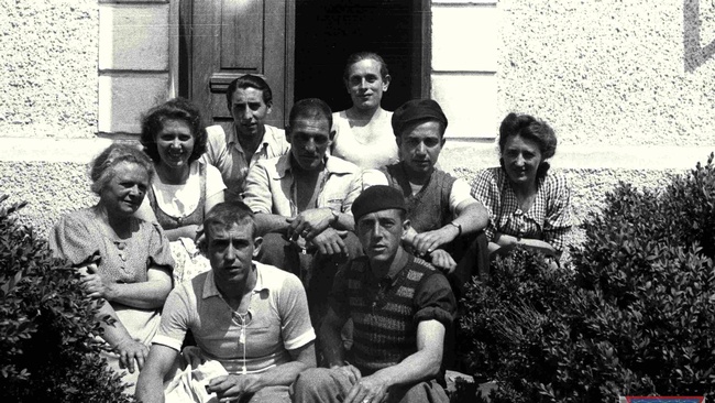 Das Schwarz-Wei&szlig;-Foto zeigt eine Gruppe von Menschen, die vor einem Haus auf der Treppe zur Eingangst&uuml;r sitzen. Ganz links sitzt Anna Pointner. Sie tr&auml;gt eine Bluse mit Kn&ouml;pfen und kurzen &Auml;rmeln. Ihre Haare sind hochgesteckt. In der Mitte der Gruppe sitzen sechs Spanier, die f&uuml;r die Kamera posieren. Einer davon l&auml;chelt, die anderen schauen ernst in die Kamera. Ein Mann ganz vorne h&auml;lt eine Zigarette in der Hand. Links und rechts der Spanier sitzt jeweils eine Tochter von Anna Pointner und l&auml;chelt in die Kamera. Im Vordergrund rahmen B&uuml;sche die Gruppe. &copy; Museu d'Hist&ograve;ria de Catalunya, Fons Amical de Mauthausen, Fotograf: Francisco Boix, Anna Pointner mit zwei T&ouml;chtern und sechs Spaniern vor ihrem Haus in Mauthausen, Mai 1945. 