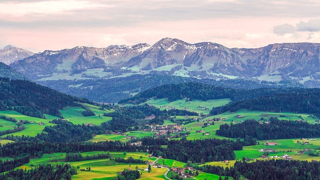 Blick auf das Dorf Krumbach: Eine Straße schlängelt sich durch die Landschaft. Zwischen gelben und hellgrünen Feldern und dunkelgrünen Waldstücken befinden sich Häusergruppen und Bauernhöfe. Im Hintergrund befindet sich eine Bergkette mit Schneeresten.  © Wikimedia, P&M Fotografie Stefan Emmer, Krumbach, 2018 (CC-BY-SA-4.0). 