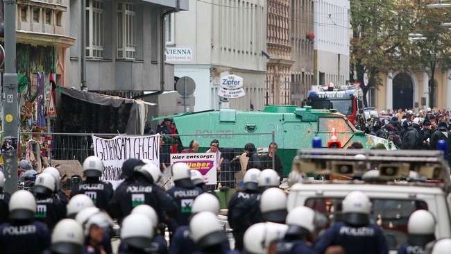 Vor dem besetzten Haus stehen viele Polizist:innen in Uniform mit weißen Helmen. An der Fassade des Hauses befinden sich Transparente und Graffitis. Auch auf einem Absperrgitter vor dem Haus sind Plakate angebracht. Im Hintergrund stehen Zuschauer:innen und weitere Polizist:innen mit Schilden.  © Wikimedia, Emanuel Sanchez de la Cerda, Räumung der Mühlfeldgasse 12, 2014 (CC-BY-2.0). 