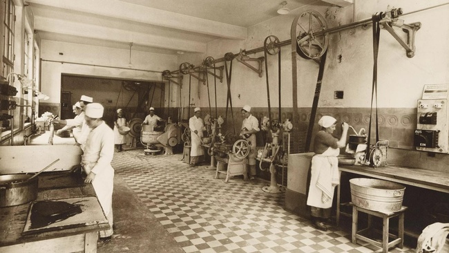 Frauen mit Schürze und Kopfbedeckung stehen an der linken und rechten Seite eines großen Raumes und kochen mit damals modernen Geräten.  © Wien Museum