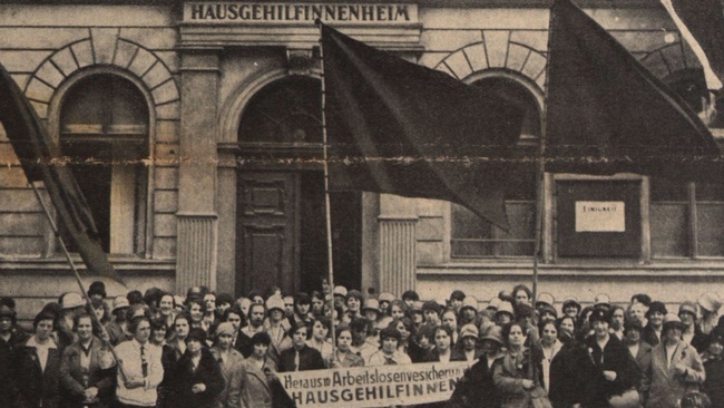 Eine große Gruppe Frauen in Festkleidung mit drei großen Fahnen und einem Banner steht vor dem Eingang zu einem Haus. Auf dem Banner ist zu lesen: Heraus mit der Arbeitslosenversicherung für Hausgehilfinnen.  © ANNO ÖNB