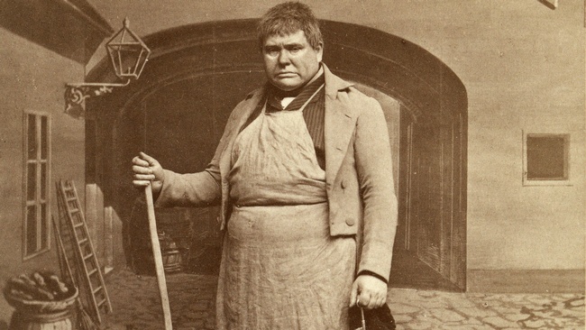 Ein Mann steht mit Besen und Schlüssel in der Hand auf einem gepflasterten Platz. Hinter ihm ist ein Tor zu sehen.  © Wien Museum
