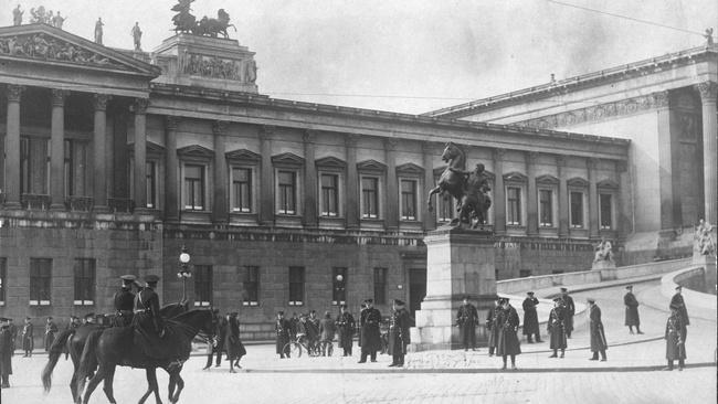 Zwei berittene Polizisten und mehrere stehende Polizisten bewachen die Rampe, die zum Parlament führt, dahinter das Parlamentsgebäude mit Säulen und Steinskulpturen am Dach.  © ÖNB Bildarchiv