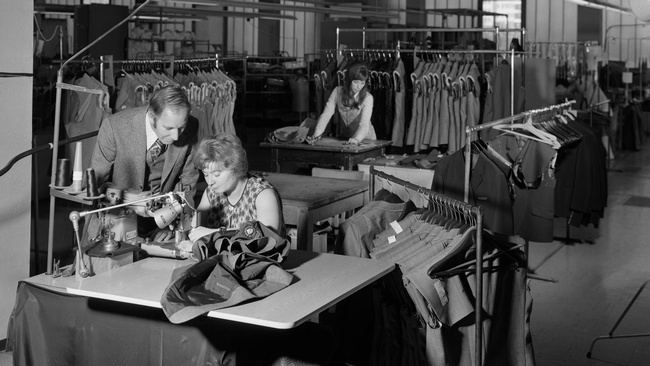 Eine Frau bedient eine Nähmaschine auf einem Schreibtisch in einer Näherei, ein Mann beugt sich über sie und sieht ihr bei der Arbeit zu. Rundherum Kleiderständer, im Hintergrund eine zweite Frau an einem Schreibtisch.  © ÖNB Bildarchiv