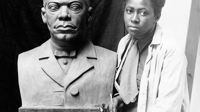 Selma Burke sitzend. Sie trägt einen weißen Mantel. In der rechten Hand hält sie ein kleines Werkzeug, möglicherweise eine schmale Spachtel. Sie sitzt Schulter an Schulter mit der Skulptur eines Mannes (Booker T. Washington). Die Skulptur steht auf einem hohen schmalen Holztisch. © Wikimedia, Photograph of Selma Burke with her portrait bust of Booker T. Washington, um 1935
