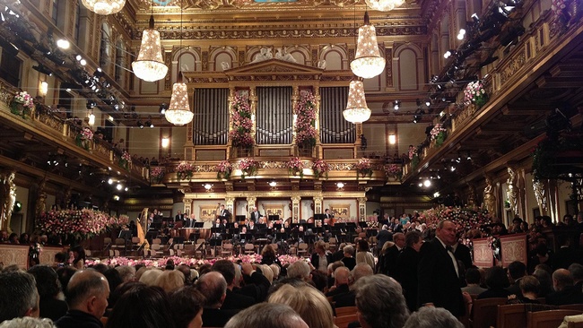 Das Bild zeigt Reihen von Zuschauer:innen beim Neujahrskonzert im großen Musikvereinssaal. Die Decke und die Logen sind mit goldenem Stuck und Malereien verziert. Im Hintergrund des Fotos befindet sich die Bühne mit den Musiker:innen. Von der Decke hängen sechs große Kronleuchter.  © Wikimedia, Michael Osmenda, Wiener Neujahrskonzert, 2013, CC-BY-2.0. 