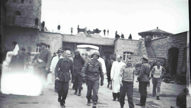 Das Schwarz-Wei&szlig;-Foto zeigt den Garagenhof des KZ Mauthausen am 5. Mai 1945. Im Hintergrund ist ein Tor zu sehen, &uuml;ber dem ein Reichsadler befestigt ist. In der Bildmitte befindet sich Albert Kosiek in Milit&auml;runiform und mit Helm, der zu Fu&szlig; den Hof durchquert. Au&szlig;erdem sind im Hof einige KZ-&Uuml;berlebende sowie Wachen der Wiener Feuerschutzpolizei in Uniform. Links im Bild befindet sich ein wei&szlig;er Fleck durch eine Besch&auml;digung der Kamera &copy; Mauthausen Memorial, Sammlung Antonio Garcia, S 4665, Fotograf: Francisco Boix, Albert Kosiek im Garagenhof, 5. Mai 1945, Signatur: 4.2.2.003.B3.