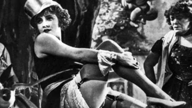 : Bild aus dem Film „Der blaue Engel“ aus dem Jahr 1930: Die Schauspielerin Marlene Dietrich posiert in der Rolle der Sängerin Lola Lola auf einem Fass. Sie trägt einen Hut, knappe Kleidung, einen Spitzenunterrock, Strumpfhalter und Stöckelschuhe. Im Hintergrund befindet sich ein Varieté-Theater mit weiteren Darstellerinnen.  © Wikimedia