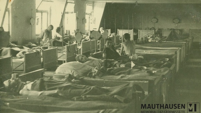 Das Schwarz-Wei&szlig;-Foto zeigt Bettenreihen in einem Innenraum. In den Betten liegen und sitzen geschw&auml;chte M&auml;nner. Zwei der ehemaligen H&auml;ftlinge schauen in die Kamera. &copy; Mauthausen Memorial, Fotoalbum Jerzy Ginter, Sanit&auml;tsbereich der US-Armee im befreiten KZ Gusen, Mai/Juni 1945, Signatur: 4.4.2.1.013.
