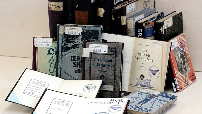 Alte Bücher mit zum Teil aufgeriebenen Kanten und Inventarnummern am Buchdeckel sind aufgereiht. Zwei Bücher im Vordergrund sind aufgeschlagen, Stempel sind sichtbar. Ein Titel lautet „Der Schilauf im Hochgebirge“, ein anderer „Wie lerne ich Schi-Laufen?“ © Marc Drews. 