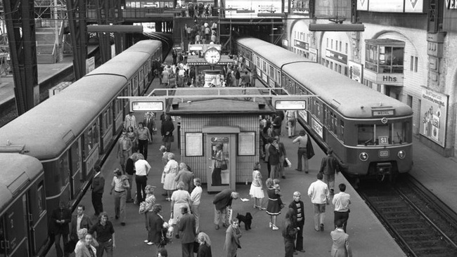 Auf dem Bahnsteig tummeln sich Menschen, rechts und links stehen auf den Gleisen Züge. In der Mitte des Bahnsteigs sind kleine Warteräume, Anzeigetafeln und eine Uhr. © Wikimedia