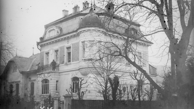 Drei Frauen, davon zwei in Arbeitskleidung mit Kleid und Schürze, stehen vor dem Eingangstor zu einer Nobelvilla mit Eckturm. Ein Kind sitzt neben ihnen auf einem Stuhl am Gehweg. Am Balkon der Villa steht ein Mann. Hinter dem Gartenzaun sind Bäume.  © ÖNB Bildarchiv