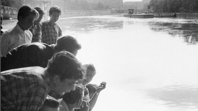 Mehrere jugendliche Burschen sitzen und stehen an einer Uferkante des Wiener Donaukanals. Im Hintergrund ist der Donaukanal von Häusern und Bäumen gesäumt. Der helle Himmel spiegelt sich im Wasser des Kanals.  © Harry Weber, ÖNB Bildarchiv. 