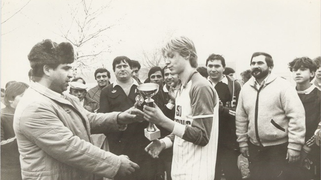 Ein Mann mit Pelzmütze überreicht einem jungen Mann im Fußballtrikot einen Pokal. Im Hintergrund stehen weitere Männer und Buben und schauen zu.  © Wien Museum, Jovan Ritopečki. 