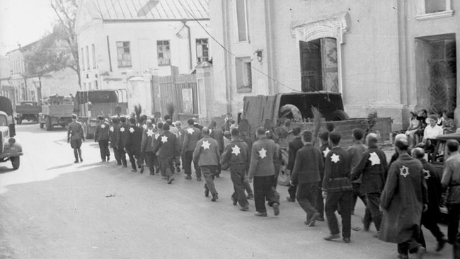 Eine Gruppe von Männern geht am Straßenrand entlang. Auf der Kleidung tragen die Männer helle Sterne. Vorne geht ein Soldat in Uniform. Rechts im Bild stehen einige Passant:innen. Die Straße ist von Häusern gesäumt.  © Wikimedia, Bundesarchiv, Bild 101I-138-1083-31, Rudolf Kessler, Kolonne von Juden mit aufgenähten Judenstern und Spaten unter Bewachung von Wehrmachtssoldaten beim Marsch durch die Stadt, CC-BY-SA 3.0. 