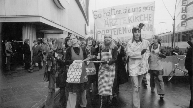 Ein Demonstrationszug, an dessen Spitze vier Frauen marschieren, eine davon mit Besen in der Hand, zwei mit Topfdeckel und Kochlöffel. Dahinter ein Banner mit der Aufschrift „So hätten sie uns gern: Ärzte, Kirche, Parteien und Männer“. Links sind Passanten vor einem Gebäude zu sehen.  © Wikimedia, Inge Werth, Frauenprotest gegen Paragraph 218, Frankfurt 1974 (CC BY-SA 4.0). 