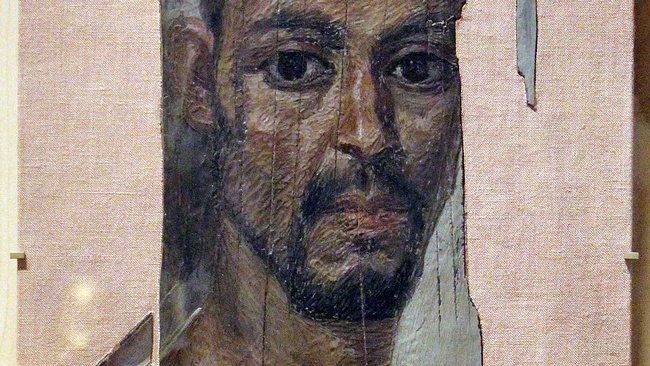 Porträt eines Mannes mit dunklen Haaren, Augen und Bart und weißer Kleidung. © Wikimedia, Mumienporträt. 