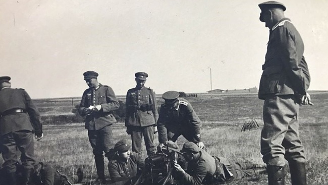 Mehrere Soldaten stehen auf einem Feld. Vier liegen am Boden und hantieren mit Kriegsgerät. Die Männer tragen Uniformen und Kappen der Wehrmacht.  © Wikimedia, Paul Calwer, Ausbildung von Wehrmachtssoldaten bei Regensburg (CC-BY-SA-4.0). 