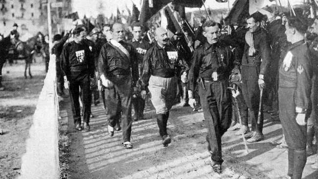 Eine Straße voller Männer in schwarzen Hemden mit Abzeichen darauf. Sie marschieren mit Fahnen, Knüppeln und Säbeln in der Hand. In der Mitte des Bildes ist Benito Mussolini zu sehen. Auch er trägt ein schwarzes Hemd und marschiert mit. © Wikimedia