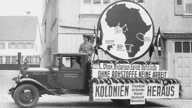 Ein altmodischer Lastwagen steht vor einem Haus. Ein Mann sitzt am Steuer. Ein zweiter Mann steht auf der Ladefläche. Er trägt einen Tropenhut und hat eine Posaune in der Hand. Der Wagen ist mit Hakenkreuzflaggen und Tannenzweigen geschmückt. Auf der Ladefläche sind außerdem drei Schilder in Form einer Treppe angeordnet: „Ohne Kolonien keine Rohstoffe”, „OHNE ROHSTOFFE KEINE ARBEIT” und „KOLONIEN HERAUS”. Am untersten ist ein zusätzliches kleineres Schild angebracht: „Frauenbund der Deutschen Kolonial-Gesellschaft Abteilung Tailfingen”. Auf dem obersten Schild steht ein rundes Holzschild mit einem Durchmesser von ungefähr 2,5 Metern. Darauf ist eine Karte von Afrika zu sehen. Togo, Kamerun, Deutsch-Ostafrika und Deutsch-Südwestafrika sind hervorgehoben. Daneben ist vergleichsweise klein das Deutsche Reich in den Grenzen von 1933 zu sehen. © Wikimedia, Werbung der Deutschen-Kolonialgesellschaft, Rückgewinnung ehemaliger Kolonien in Afrika, 1933.