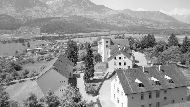 Im Vordergrund sind in einer Gruppe vier Heimgebäude umgeben von Bäumen zu sehen. Der Blick öffnet sich auf ein weites Tal mit Feldern und Wald. Berge im Hintergrund schließen es ab.  © Wikimedia, Landeserziehungsanstalt Jagdberg - Schlins / Vorarlberg, 1961.