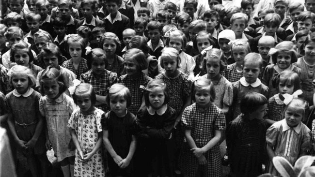 Zahlreiche Kinder stehen in einer Gruppe beieinander und schauen zur fotografierenden Person hoch. Vorne stehen in mehreren Reihen Mädchen in Kleidern, dahinter Buben. In den hintersten Reihen stehen Erwachsene in weißen Kitteln und mit weißen Hauben.  © ÖNB Bildarchiv, Kinder am Steinhof nach ihrer Befreiung 1945. 