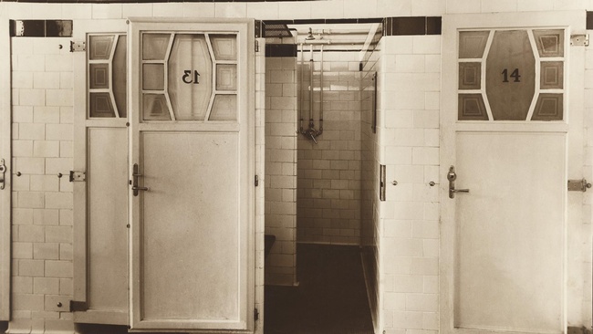 Das Foto zeigt wei&szlig; geflieste W&auml;nde und G&auml;nge mit wei&szlig;en T&uuml;ren, die zu den Duschkabinen f&uuml;hren. In den T&uuml;ren befindet sich oben ein Fenster mit einer Nummer, einmal &bdquo;13&ldquo; und einmal &bdquo;14&ldquo;. Der Boden ist dunkel.  &copy; Wien Museum, St&auml;dtisches Volksbad in der Ratschkygasse, 1926. 