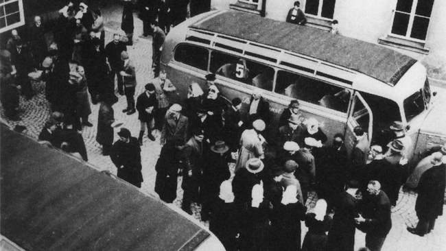 Auf einem gepflasterten Platz umgeben von Gebäuden stehen zwei Busse. Zwischen den leeren Bussen stehen mehrere Dutzend Menschen. © Wikimedia, Pressestelle der Diakonie Neuendettelsau, Aktion T4 (Diakonie Neuendettelsau), 1941