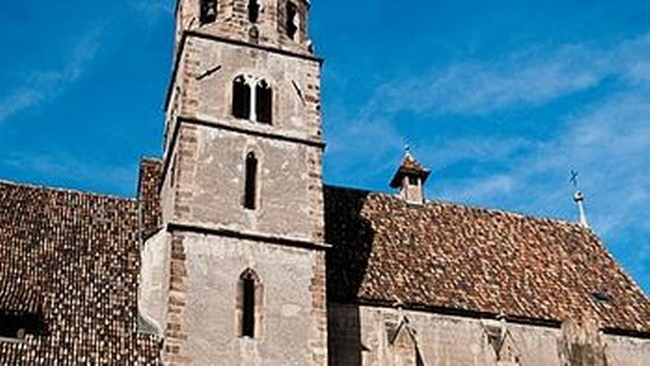 Die Kirche des Franziskanerklosters besteht aus graubraunen Steinmauern und einem Schindeldach mit verschiedenfarbigen Schindeln. Mittig im Foto befindet sich ein Kirchturm mit schmalen, spitz zulaufenden Fenstern. Die Spitze des Kirchturms besteht aus einem hellen Dach. Auch die Fenster des Kirchenschiffs laufen spitz zu und sind teilweise mit verschnörkelten Elementen verziert.  © Wikimedia, Bigmike.it, Franziskanerkloster in Bozen (CC-BY-SA-3.0). 