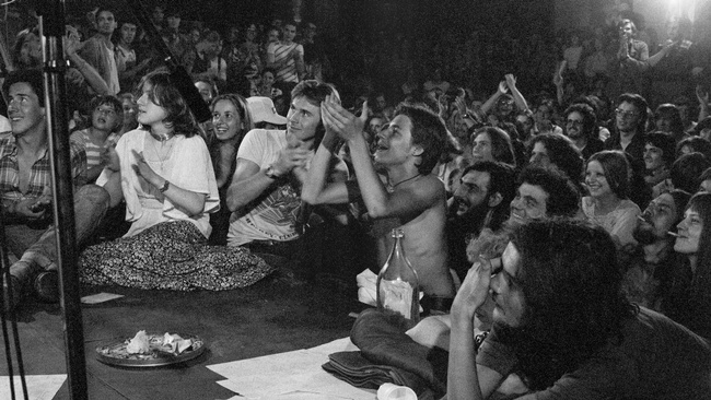  An der Ecke einer Bühne, auf der ein Mikrophon steht, sitzen und stehen Jugendliche. Sie lachen und klatschen. Der Saal ist voller junger Leute.  © Heinz Riedler, Wien Museum. 