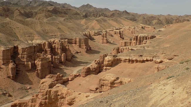 Das Foto zeigt einen Canyon mit Bergen im Hintergrund vor einem graublauen Himmel. Die Erde und das Gestein des Canyons sind orange.  © Wikimedia, Anna Dobrynina, Canyon in Kazakhstan, 2017 (CC-BY-3.0).  