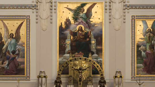 Drei prächtige, goldene Bilder hängen über einem Altar. Auf dem linken Bild sind zwei Engel zu sehen. Das mittige Bild zeigt Jesus Christus auf einem Thron. Ein betender Mann lehnt sich auf ihn. Über den beiden schwebt ein Engel, der eine Feder in der Hand hält. Im rechten Bild steht Karl Lueger zwischen einem Engel und einer knienden Frau. Die Frau hält ein Modell der Karl-Borromäus-Kirche in den Händen. © Wikimedia, Presbyterium der Karl-Borromäus-Kirche am Wiener Zentralfriedhof, 2014.