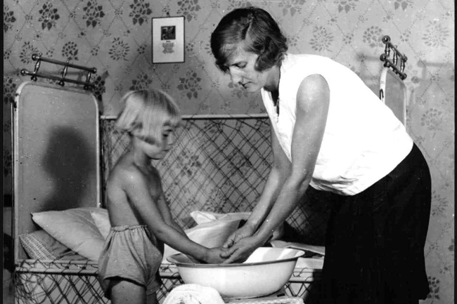 Eine Frau mit Bubikopf, Bluse und Rock wäscht einem Mädchen die Hände in einer Waschschüssel. Hinter ihnen ein kleines Bett vor einer tapezierten Wand.  © ÖNB Bildarchiv