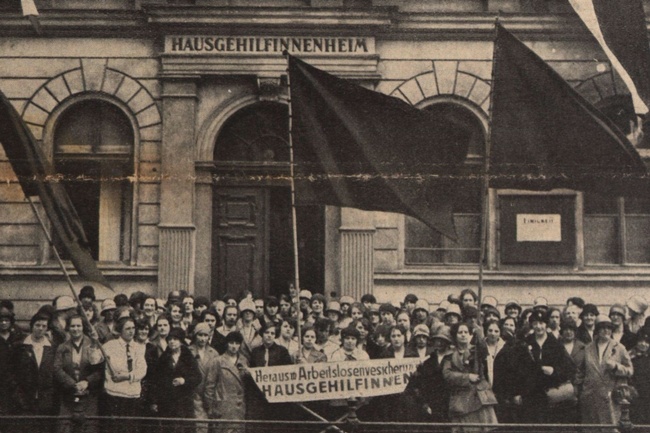 Eine große Gruppe Frauen in Festkleidung mit drei großen Fahnen und einem Banner steht vor dem Eingang zu einem Haus. Auf dem Banner ist zu lesen: Heraus mit der Arbeitslosenversicherung für Hausgehilfinnen.  © ANNO ÖNB