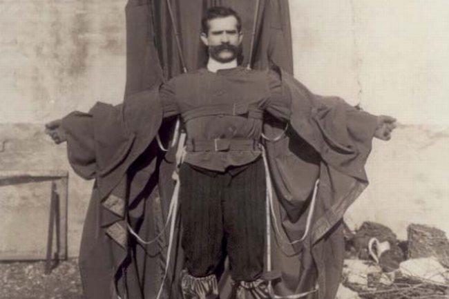 : Ein Mann mit bauschigem Schnauzer trägt dünne Gummistiefel und einen Fallschirmanzug, der mit Bändern und Schnüren an seinem Körper befestigt ist. Seine Arme streckt er seitlich aus. Er steht breitbeinig vor einer Hausmauer im Freien. © Wikimedia