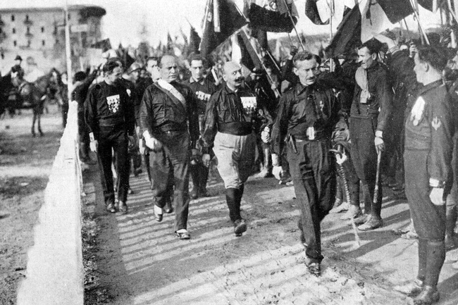 Eine Straße voller Männer in schwarzen Hemden mit Abzeichen darauf. Sie marschieren mit Fahnen, Knüppeln und Säbeln in der Hand. In der Mitte des Bildes ist Benito Mussolini zu sehen. Auch er trägt ein schwarzes Hemd und marschiert mit. © Wikimedia