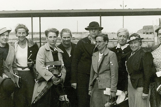 Eine Gruppe Frauen in festlicher Kleidung, einige mit Hut und Blazer, stehen nebeneinander und blicken in die Kamera. Dahinter ein Bahnsteig und eine Wiese.  © Wikimedia