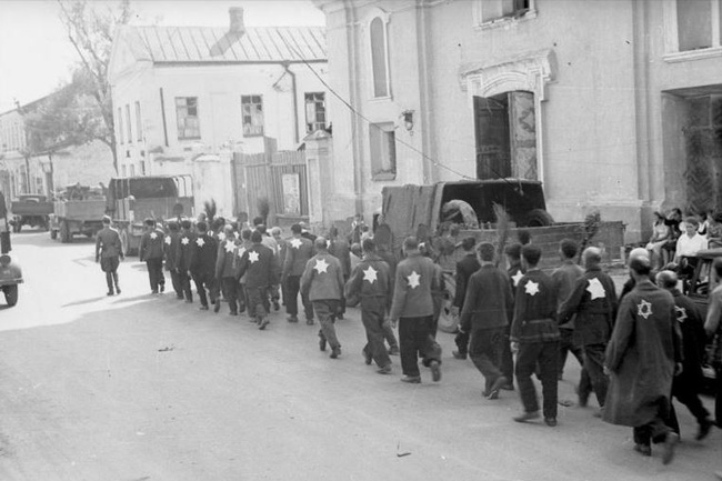 Eine Gruppe von Männern geht am Straßenrand entlang. Auf der Kleidung tragen die Männer helle Sterne. Vorne geht ein Soldat in Uniform. Rechts im Bild stehen einige Passant:innen. Die Straße ist von Häusern gesäumt.  © Wikimedia, Bundesarchiv, Bild 101I-138-1083-31, Rudolf Kessler, Kolonne von Juden mit aufgenähten Judenstern und Spaten unter Bewachung von Wehrmachtssoldaten beim Marsch durch die Stadt, CC-BY-SA 3.0. 