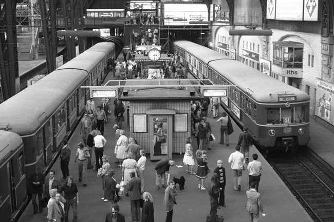Auf dem Bahnsteig tummeln sich Menschen, rechts und links stehen auf den Gleisen Züge. In der Mitte des Bahnsteigs sind kleine Warteräume, Anzeigetafeln und eine Uhr. © Wikimedia